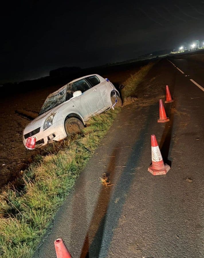 A white car in a ditch at night with cones deployed on the carriageway